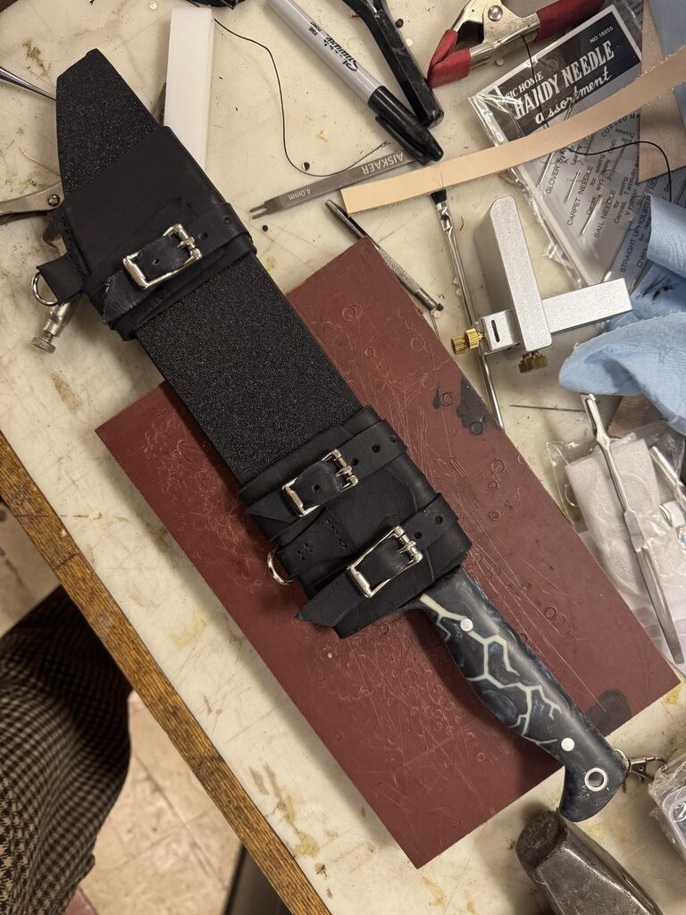 Large Sabatier-style knife secured in a black post-apocalyptic scabbard on a leatherworking bench